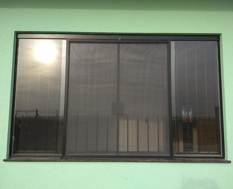 Janela de correr com perfil preto e grade, com tela mosquiteira de duas abas no vão central, vista de frente em parede verde