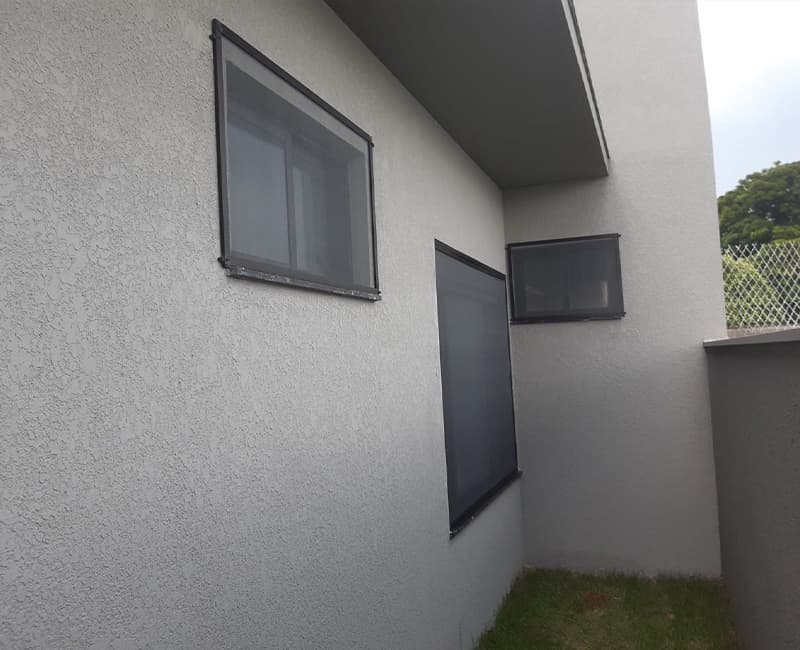 Três janelas pequenas de casa com tela mosquiteira de perfil preto instalada pelo lado de fora, em parede texturizada cinza