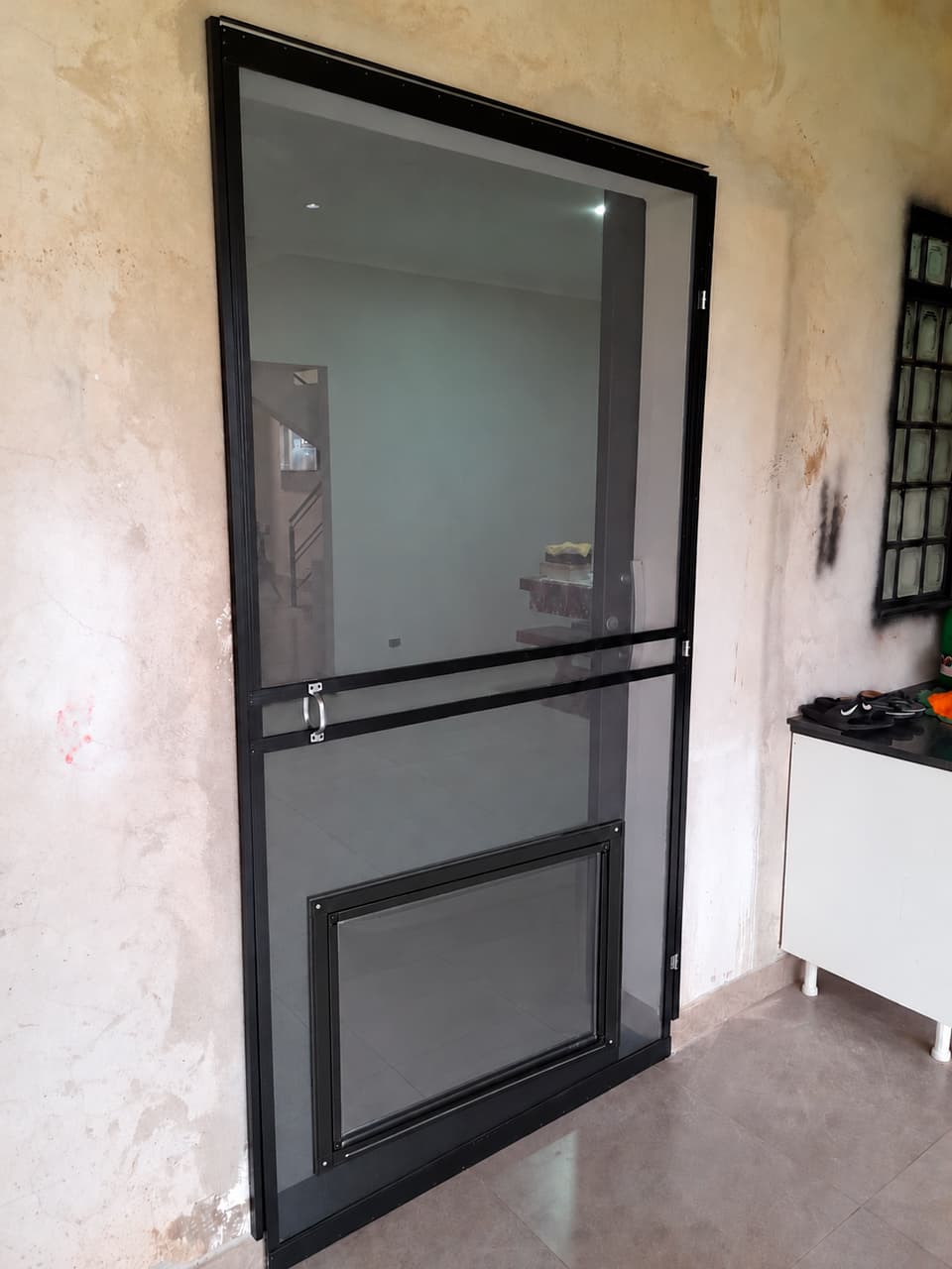 Porta de tela mosquiteira com perfil preto, puxador e passagem para pets na parte inferior, instalada em residência